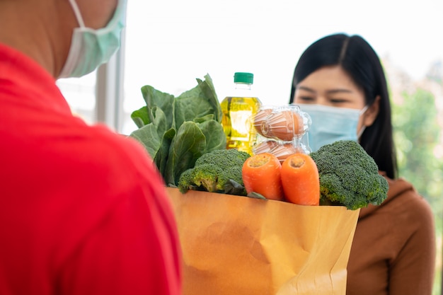 Repartidor asiático del supermercado con una máscara facial y sosteniendo una bolsa de comida fresca para dar a los clientes en casa.