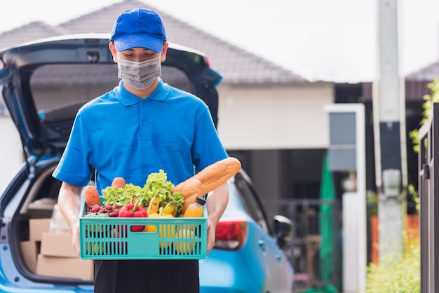 El repartidor asiático de la tienda de comestibles con uniforme azul y mascarilla protege la entrega de vegetales frescos en una caja de plástico en la puerta de la casa después del brote de coronavirus, de vuelta al nuevo concepto normal