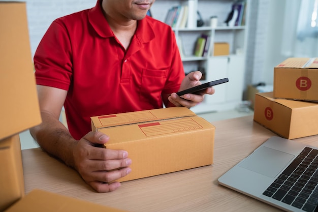 Repartidor asiático trabajador sonriendo usando código de barras de escaneo móvil para cajas para enviar o transportar paquetes por correo Usando uniforme rojo trabajo en casa Concepto de transporte de entrega