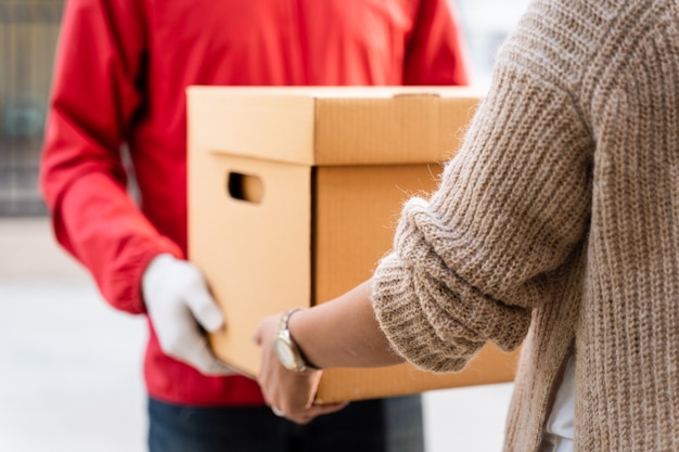 Un repartidor asiático con uniforme rojo entrega un paquete a una clienta frente a la casa. Un cartero y un servicio de entrega urgente entregan paquetes durante la pandemia de covid19.