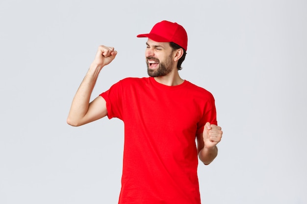 Repartidor barbudo joven alegre y despreocupado con gorra de uniforme rojo y camiseta cantando gritando sí un ...