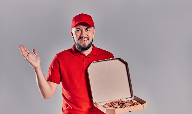 Repartidor barbudo joven en uniforme rojo con caja de pizza aislada sobre fondo gris.