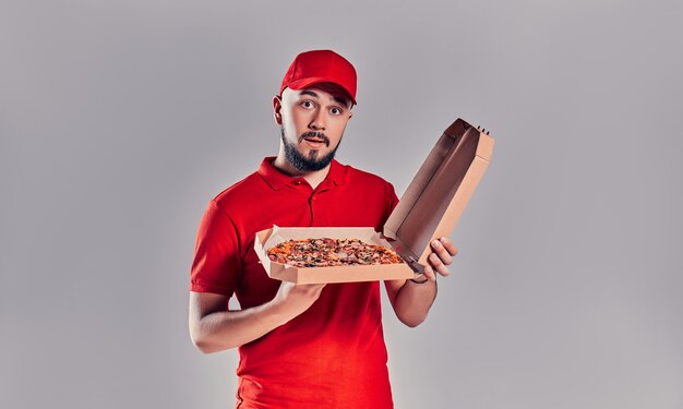 Repartidor barbudo joven en uniforme rojo con caja de pizza aislada sobre fondo gris.