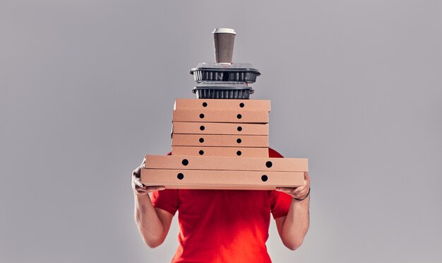 Repartidor barbudo joven en uniforme rojo con cajas de pizza, lonchera y café aislado sobre fondo gris.