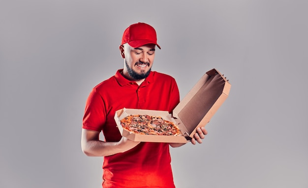 Repartidor barbudo joven en uniforme rojo mira con disgusto a la caja de pizza aislada sobre fondo gris. Comida insípida en mal estado.