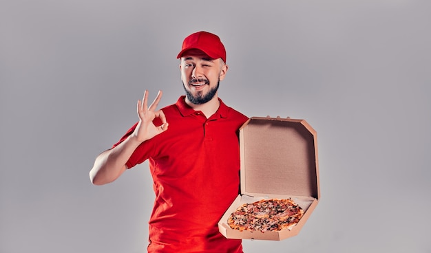 Foto repartidor barbudo joven con un uniforme rojo muestra el gesto de que todo está bien y sostiene una caja de pizza aislada sobre fondo gris.