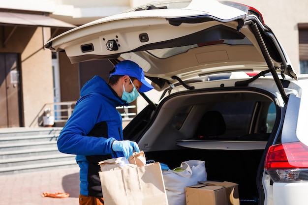 Repartidor con bolsa de papel con comida cerca del coche, repartidor de comida en máscara protectora