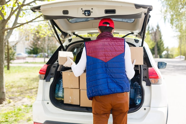 Repartidor con bolsa de papel con comida cerca del coche, repartidor de comida en máscara protectora