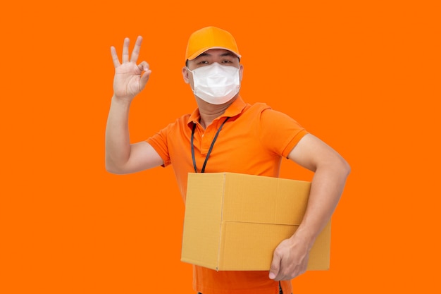 Repartidor con caja de paquetería y con máscara protectora para prevenir el virus Covid-19 y mostrando bien aislado en la pared naranja, envío de compras en línea y concepto de servicio de entrega rápida