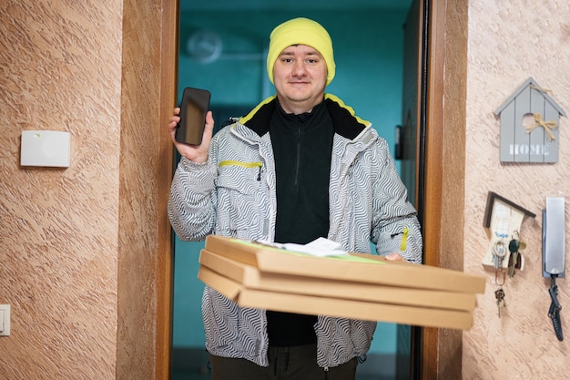 Repartidor con cajas de cartón de pizza Mensajero con sombrero de color verde con teléfono móvil de pie contra la puerta de la casa residencial