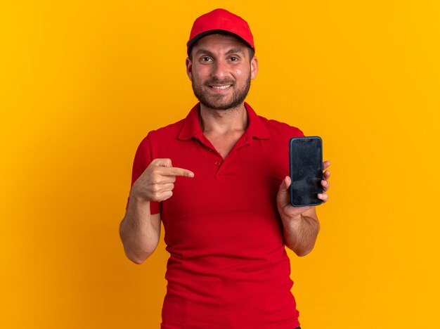 Repartidor caucásico joven sonriente en uniforme rojo y gorra mirando a cámara mostrando teléfono móvil apuntando a él aislado en la pared naranja