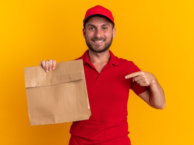 Repartidor caucásico joven sonriente en uniforme rojo y gorra que muestra el paquete de papel apuntando hacia él mirando a la cámara aislada en la pared naranja