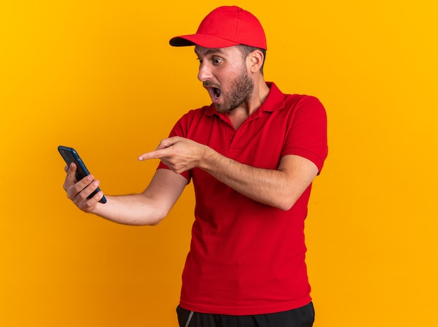 Repartidor caucásico joven sorprendido en uniforme rojo y gorra sosteniendo mirando y apuntando al teléfono móvil aislado en la pared naranja