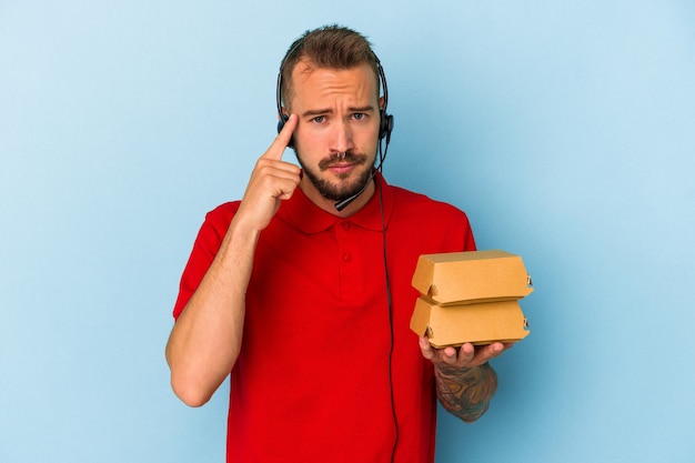 Repartidor caucásico joven con tatuajes sosteniendo hamburguesas aisladas sobre fondo azul apuntando la sien con el dedo, pensando, centrado en una tarea.