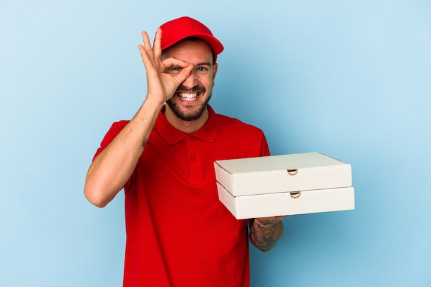 Repartidor caucásico joven con tatuajes sosteniendo pizzas aisladas sobre fondo azul emocionado manteniendo el gesto ok en el ojo.