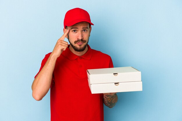 Repartidor caucásico joven con tatuajes sosteniendo pizzas aisladas sobre fondo azul señalando la sien con el dedo, pensando, centrado en una tarea.