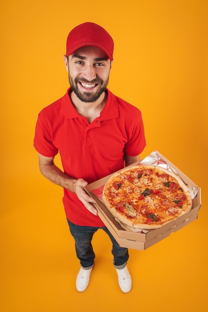 Repartidor caucásico de longitud completa en uniforme rojo sonriendo y sosteniendo una caja de pizza aislada sobre amarillo