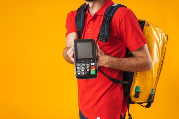 Repartidor caucásico recortada en uniforme rojo sosteniendo terminal de pago mientras lleva mochila con comida para llevar aislado sobre amarillo