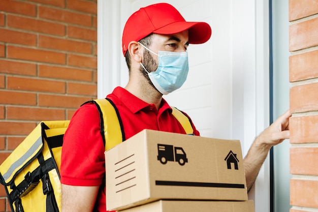 Foto repartidor de compras en uniforme rojo con mascarilla protectora tocando el timbre de la casa para entregar el paquete concepto de servicio de entrega y pequeña empresa