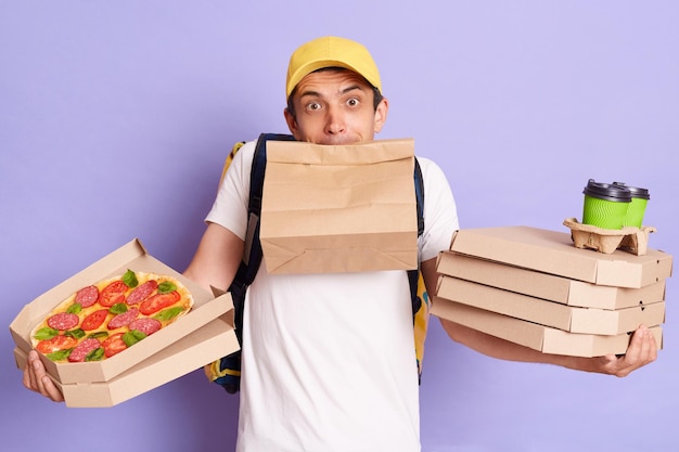 Un repartidor confundido con camiseta y gorra sosteniendo cajas de pizza y café para llevar de pie con un paquete de papel en la boca parece indefenso con muchos pedidos posando aislados sobre un fondo morado