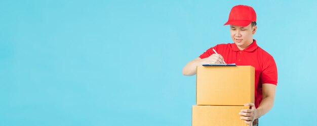 Repartidor feliz asiático vistiendo una camisa roja escribiendo bloc de notas mientras sostiene cajas de paquetes de papel