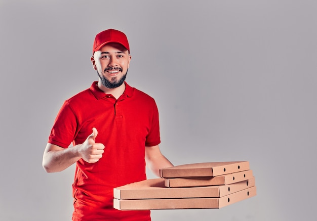 Repartidor con gorra roja, camiseta dando cajas de pizza de pedido de comida aisladas sobre fondo gris. Mensajero de pizzero empleado masculino sosteniendo pizza en el espacio vacío de la copia de la caja plana de cartón en blanco. Concepto de servicio