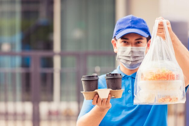 Repartidor joven asiático con uniforme azul con mascarilla haciendo servicio de comestibles dando cajas de comida de arroz bolsas de plástico y café en la casa bajo el coronavirus pandémico, de vuelta al nuevo concepto normal