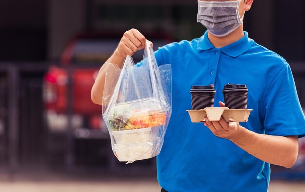 Repartidor joven asiático con uniforme azul con mascarilla haciendo servicio de comestibles dando cajas de comida de arroz bolsas de plástico y café en la casa bajo el coronavirus pandémico, de vuelta al nuevo concepto normal