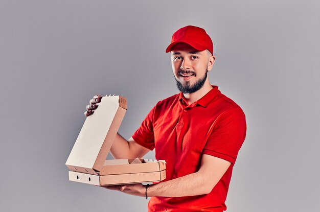 Repartidor joven barbudo en camiseta roja y gorra con cajas de pizza aisladas sobre fondo gris. Entrega rápida a domicilio.