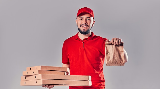 Repartidor joven barbudo en camiseta roja y gorra con cajas de pizza y bolsa de papel de comida rápida aislada sobre fondo gris. Entrega rápida a domicilio.