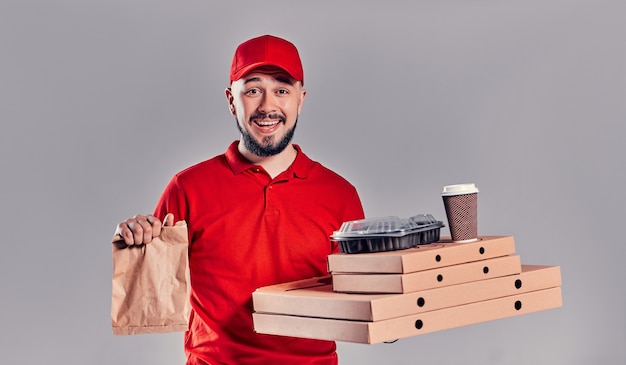 Repartidor joven barbudo en camiseta roja y gorra con cajas de pizza y bolsa de papel de comida rápida y tazas de café aisladas sobre fondo gris. Entrega rápida a domicilio.