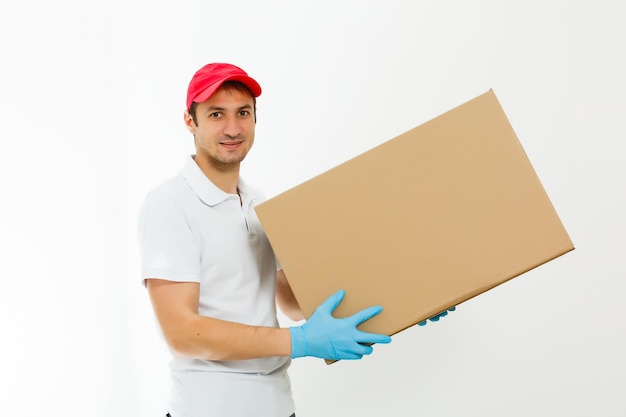 Repartidor joven sonriente sosteniendo y llevando una caja de cartón aislado sobre fondo blanco.