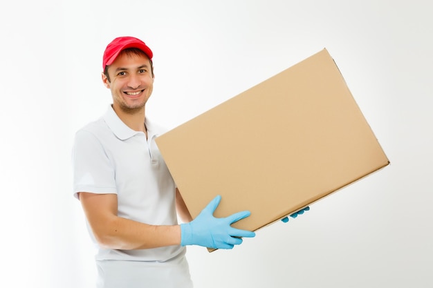 Repartidor joven sonriente sosteniendo y llevando una caja de cartón aislado sobre fondo blanco.