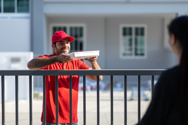 Repartidor joven sonriente en uniforme rojo sosteniendo una caja para dar a cliente hermosa mujer en frente de la casa.