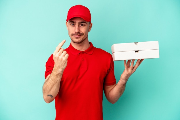 Repartidor joven tomando pizzas aisladas en azul bakcground apuntando con el dedo como si invitara a acercarse.