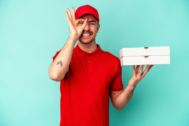 Repartidor joven tomando pizzas aisladas en azul bakcground emocionado manteniendo el gesto ok en el ojo.