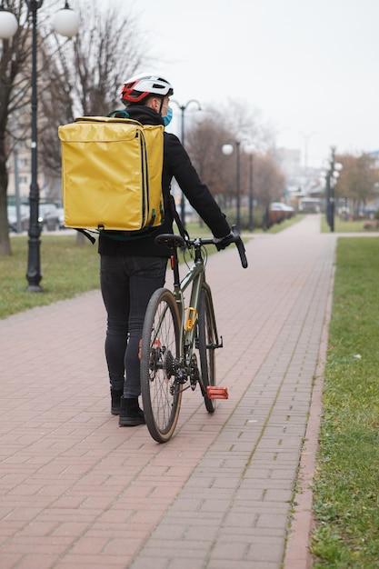 Repartidor con máscara médica y mochila térmica, caminando en la ciudad después de andar en bicicleta