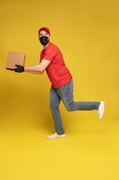 Repartidor en máscara protectora y guantes con caja en estudio aislado en pared amarilla