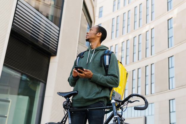 Un repartidor con una mochila amarilla está buscando el edificio correcto Un hombre afroamericano en el contexto de los edificios modernos de la ciudad