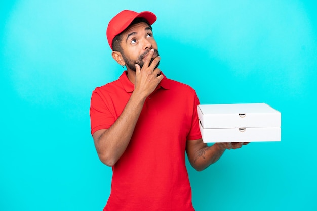Repartidor de pizza brasileño con uniforme de trabajo recogiendo cajas de pizza aisladas de fondo azul con dudas mientras mira hacia arriba