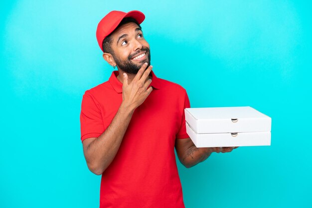 Repartidor de pizza brasileño con uniforme de trabajo recogiendo cajas de pizza aisladas de fondo azul mirando hacia arriba mientras sonríe