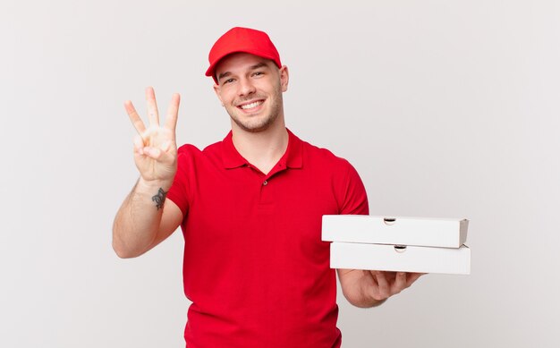 Repartidor de pizza hombre sonriendo y mirando amigable, mostrando el número tres o tercero con la mano hacia adelante, contando hacia atrás