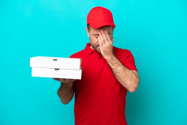 Repartidor de pizza con uniforme de trabajo recogiendo cajas de pizza aisladas de fondo azul con expresión cansada y enferma