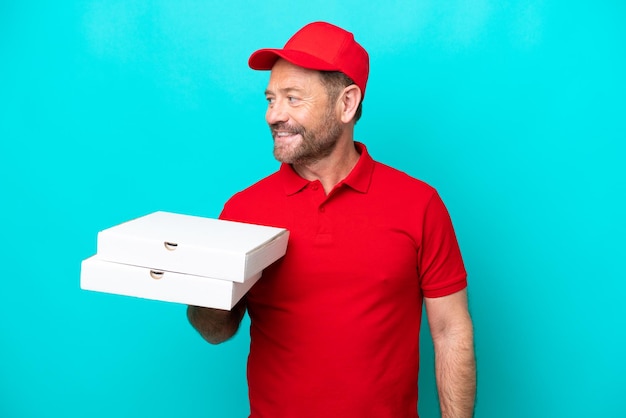 Repartidor de pizza con uniforme de trabajo recogiendo cajas de pizza aisladas sobre fondo azul mirando hacia el lado