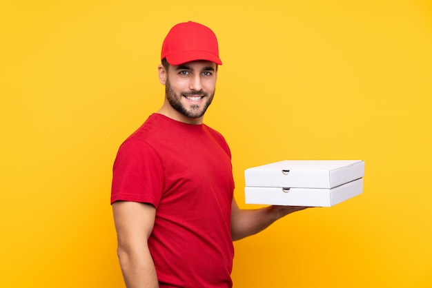 Repartidor de pizza con uniforme de trabajo recogiendo cajas de pizza sobre pared amarilla aislada sonriendo mucho