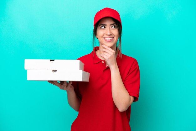 Repartidor de pizzas con uniforme de trabajo recogiendo cajas de pizza aisladas de fondo azul pensando en una idea mientras mira hacia arriba