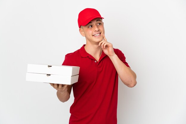 Repartidor de pizzas con uniforme de trabajo recogiendo cajas de pizza aisladas en la pared blanca pensando en una idea mientras mira hacia arriba