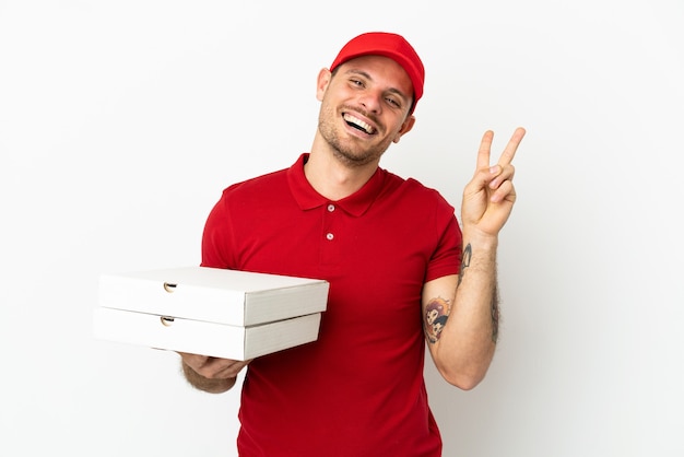 Repartidor de pizzas con uniforme de trabajo recogiendo cajas de pizza sobre una pared blanca aislada sonriendo y mostrando el signo de la victoria