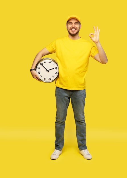 Repartidor en ropa de trabajo amarilla mantenga reloj aislado en retrato de estudio de fondo amarillo.