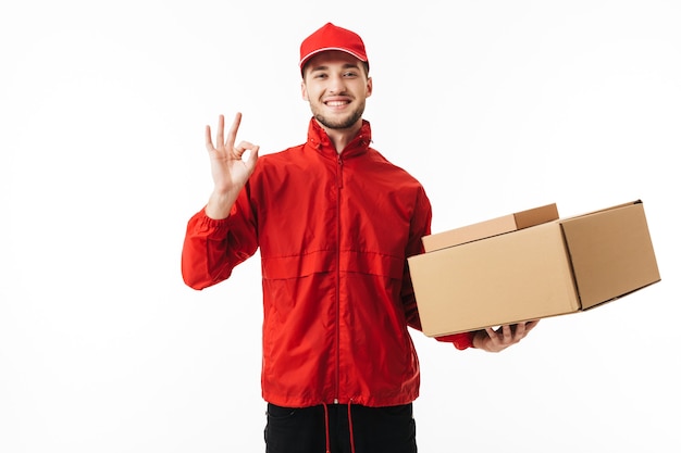 Foto repartidor sonriente joven con gorra roja y chaqueta sosteniendo cajas en la mano mostrando alegremente gesto ok